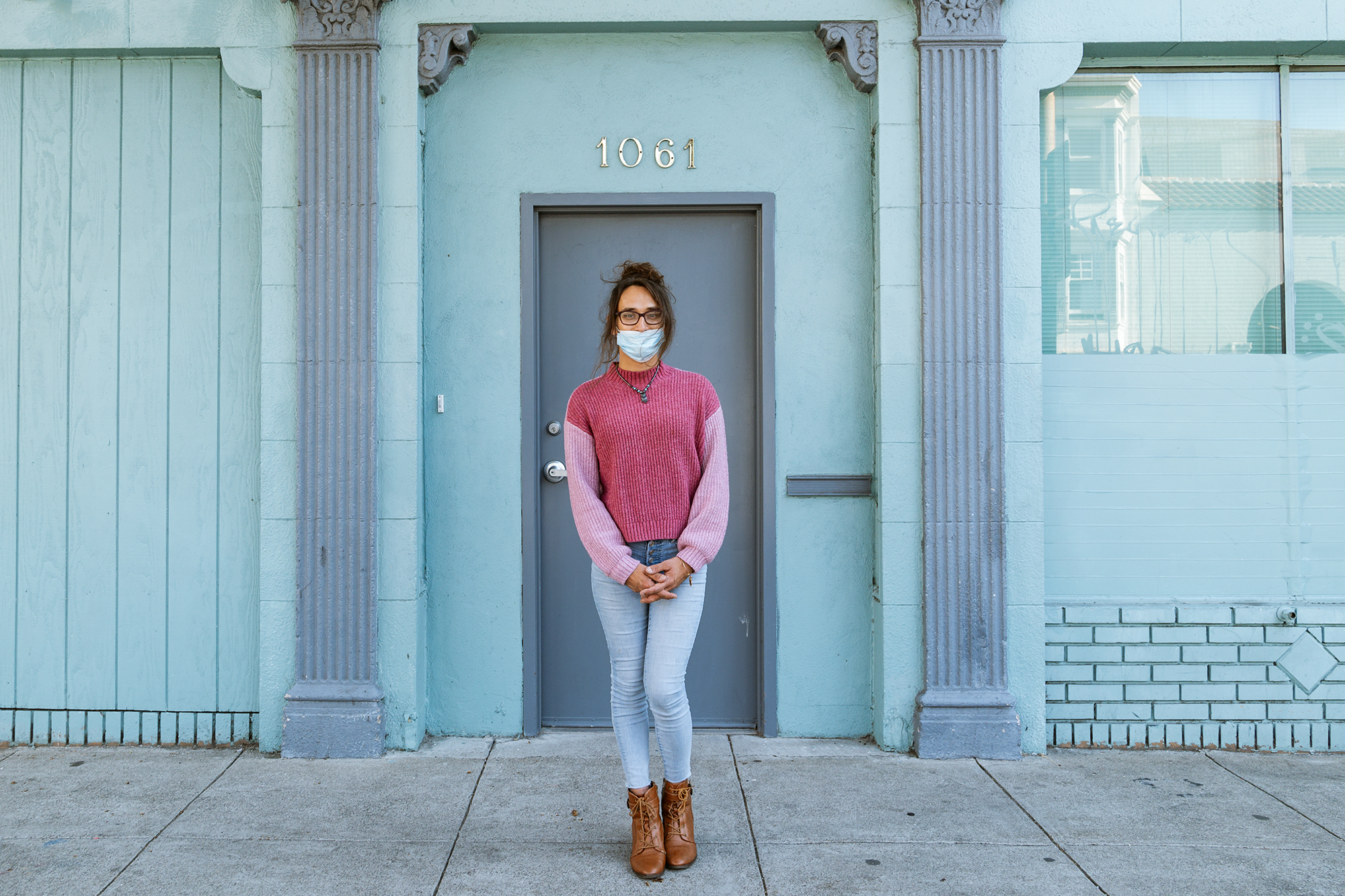 A person stands in front of an apartment building. They wear a face mask and have glasses.