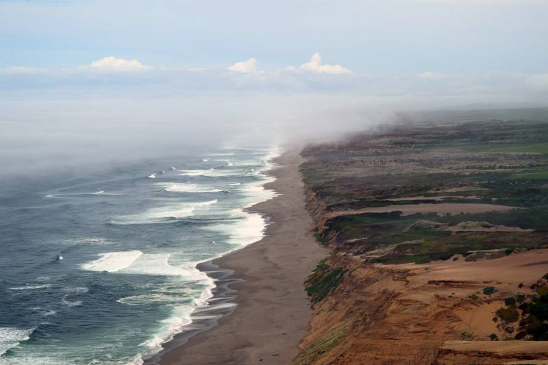 Aerial view of the California coast. On the left side of the image, the ocean laps against the shore, on the right side, plant covered sand dunes rise from the beach. Fog rolls in.