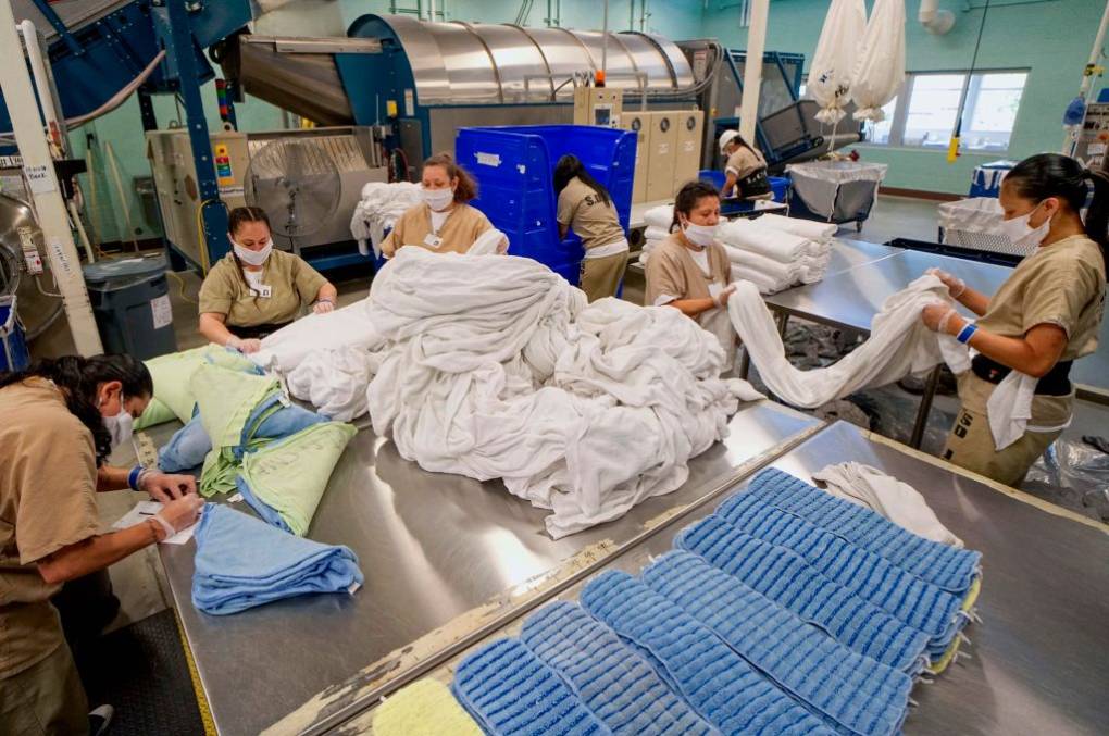 A group of female workers, in prison uniforms, fold sheets in an industrial laundry facility.