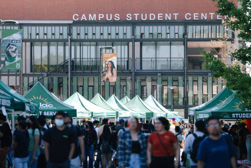 A crowd of people are seen walking around outside on a campus near green tents with a building in the background that says "Campus Student Center."