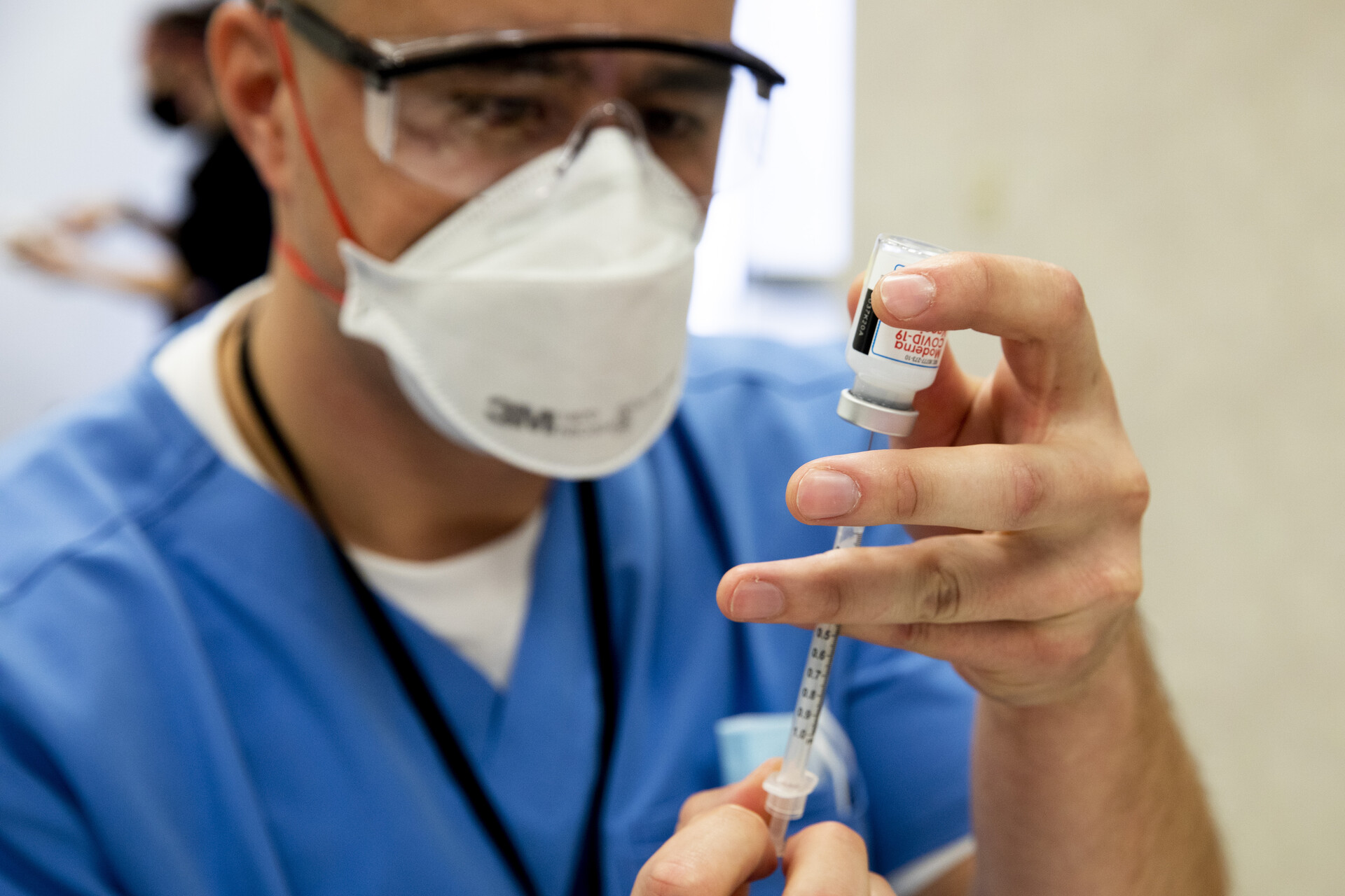 A health care worker wearing blue scrubs and a white face mask holds a vial of COVID vaccine.