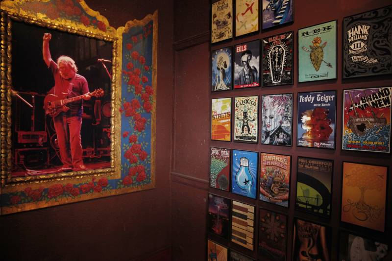A display of concert posters. On one wall are numerous framed posters for different musicians. On another wall, a large framed portrait of Grateful Dead guitarist, Jerry Garcia is visible. Garcia is playing a guitar.