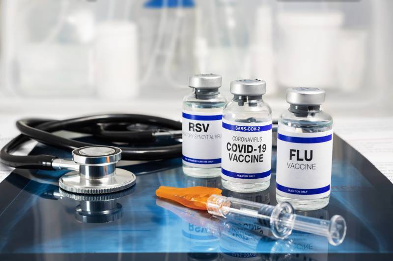 Three vaccine bottles - labeled 'RSV,' 'COVID-19' and 'Flu' sit next to a syringe and stethoscope on a medical table.
