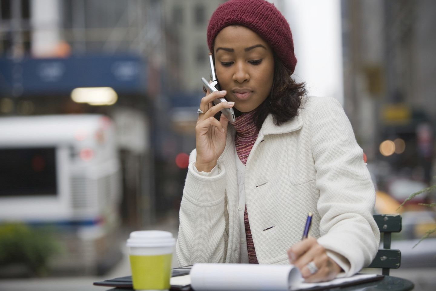 A person in a white coat and red beret sits at an outdoor table on the phone, writing on a notepad.