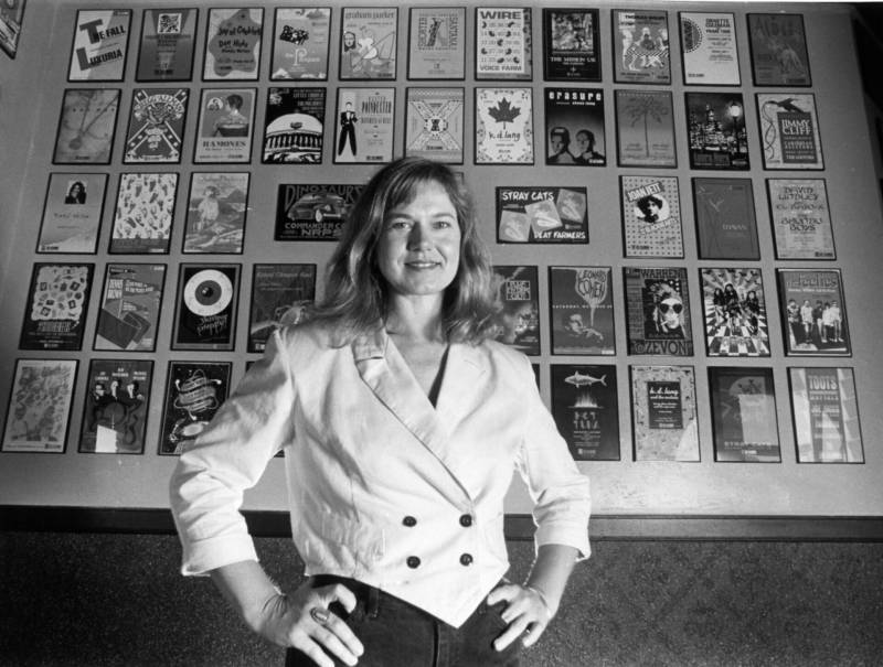 Black and white image of a woman wearing a white blazer, standing with her hands on her hips. Behind her is a wall filled with dozens of framed concert posters. 