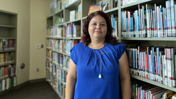 A woman wearing a blue shirt stands by a bookshelf in a library.