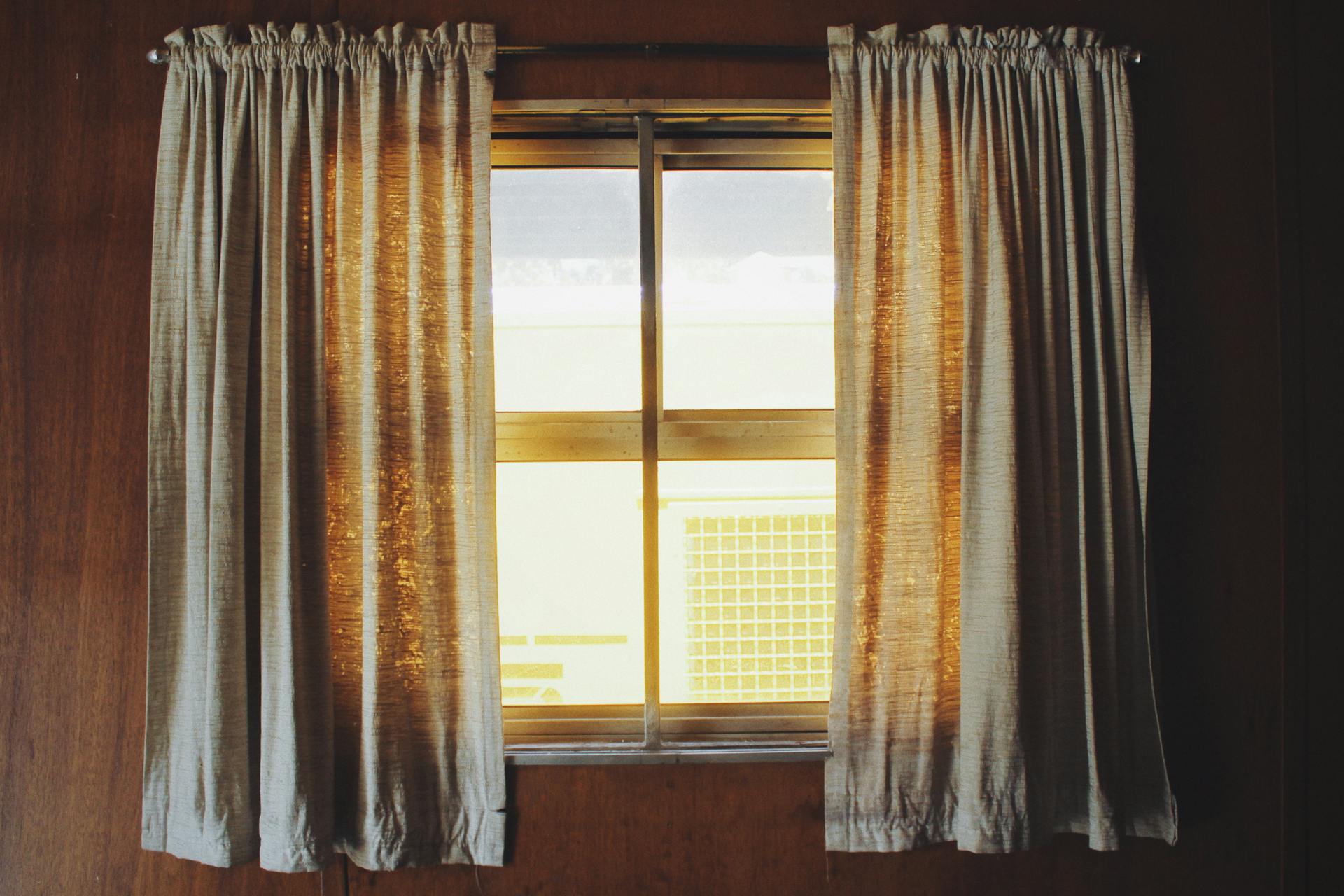 An image of a window with short grey curtains set into brown walls. There is soft yellow light coming through.