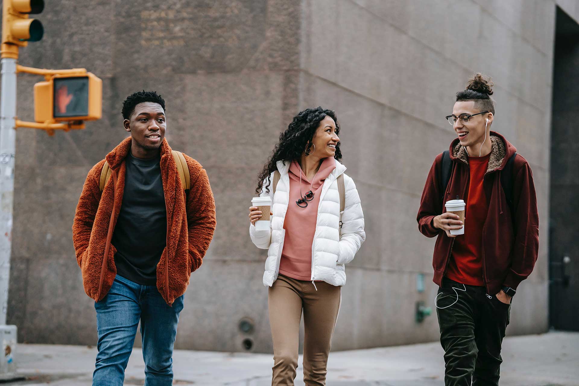 Three young people walk together on what looks like a city street, dressed warmly and smiling at each other.