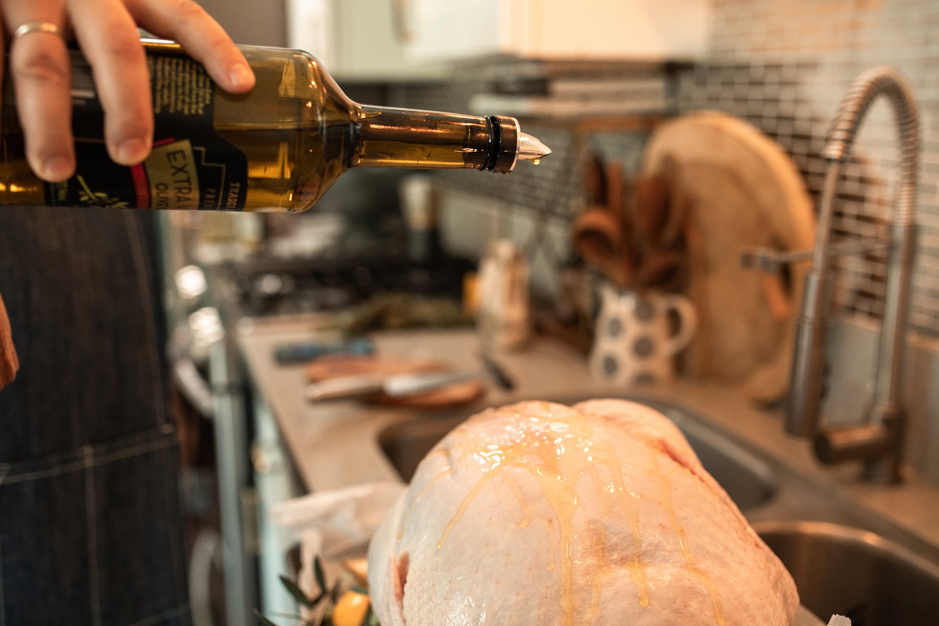 A hand holds a bottle of olive oil, drizzling the oil over a raw turkey against the backdrop of a kitchen