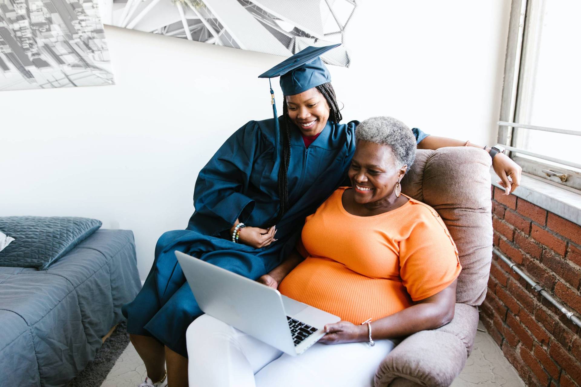 Two people, one older and one younger, sit on a stuffed seat looking at a laptop. The younger person is wearing college graduation clothes.
