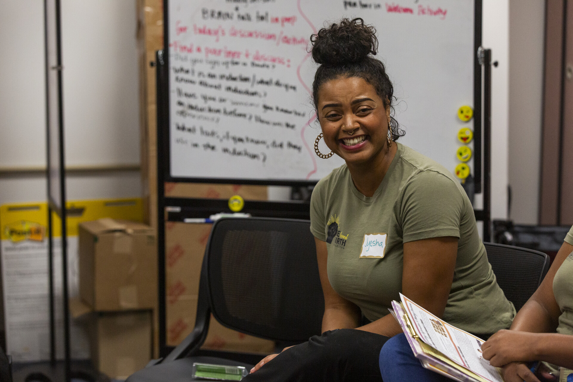 An African American woman smiles as she sits in front of a whiteboard with writing on it.