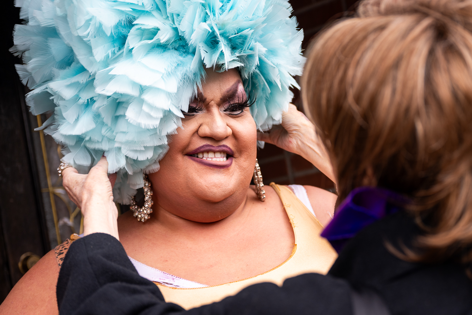 A drag artist is given a blue wig as they smile while another drag artist puts it on.