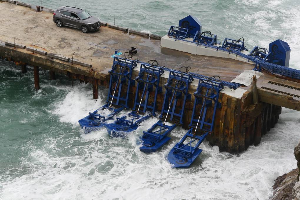 Ocean waters surround a cement platform where a vehicle is parked. On either side of the platform is equipment that aims to capture ocean waves as energy.