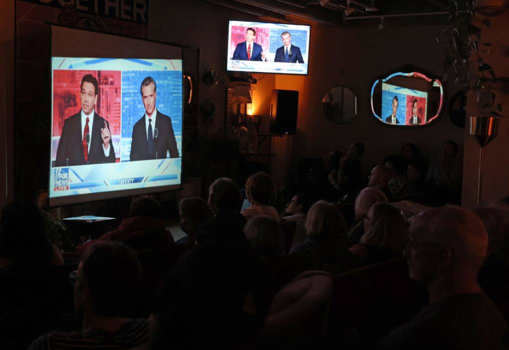 A dim-lit room full of people watching a TV screen with two men.