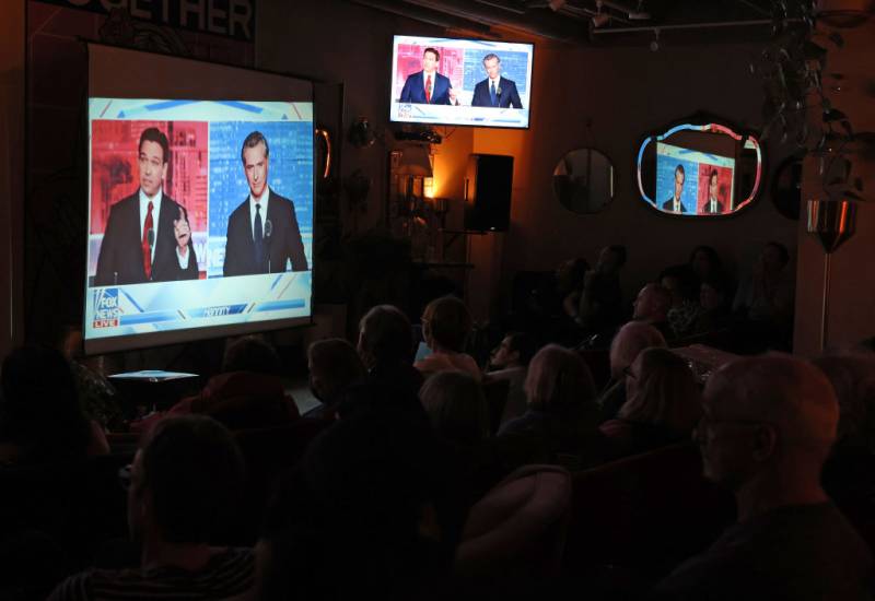 A dim-lit room full of people watching a TV screen with two men.