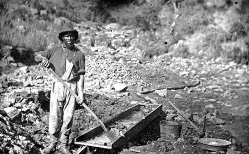 A black-and-white photo of a Black miner, holding a shovel, and standing next to a sluice.