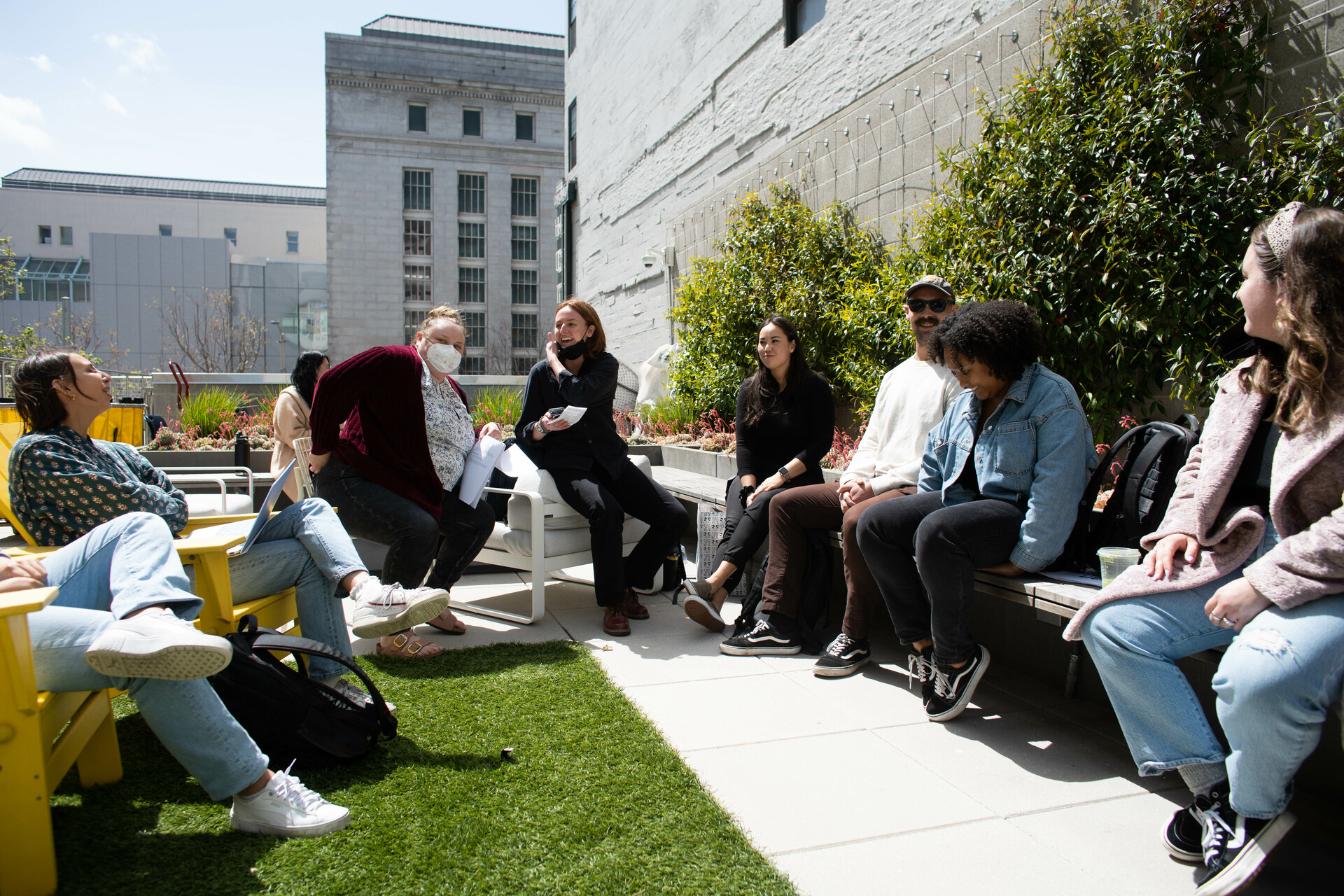 Students seated in a circle while in discussion, outside in a courtyard with buildings behind them and a little lawn underneath them.