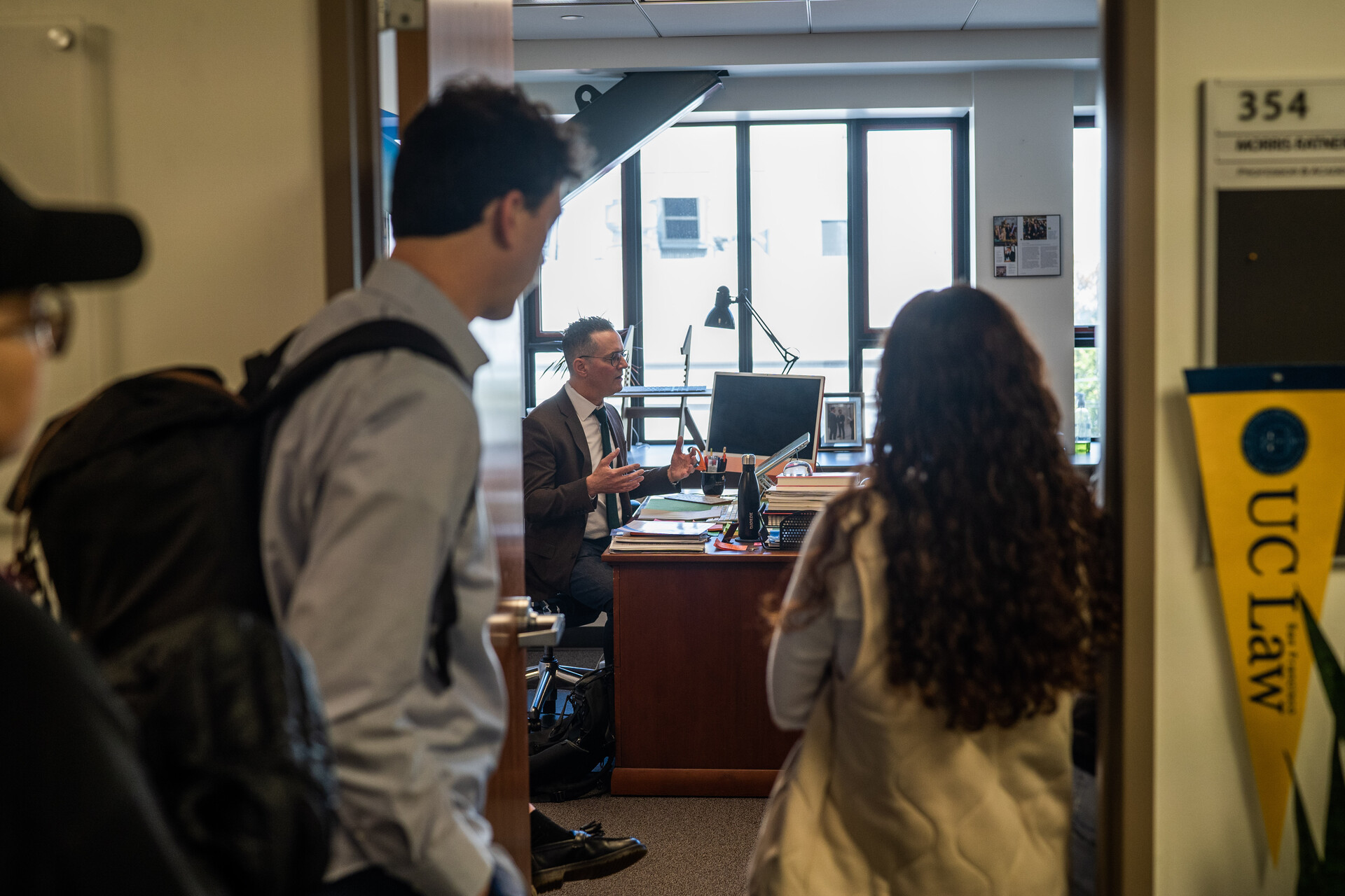 Two students, one male, the other female, stand on either side of a door with their backs to the camera, through the door we see a white man in a brown suit sitting behind a desk explaining something.
