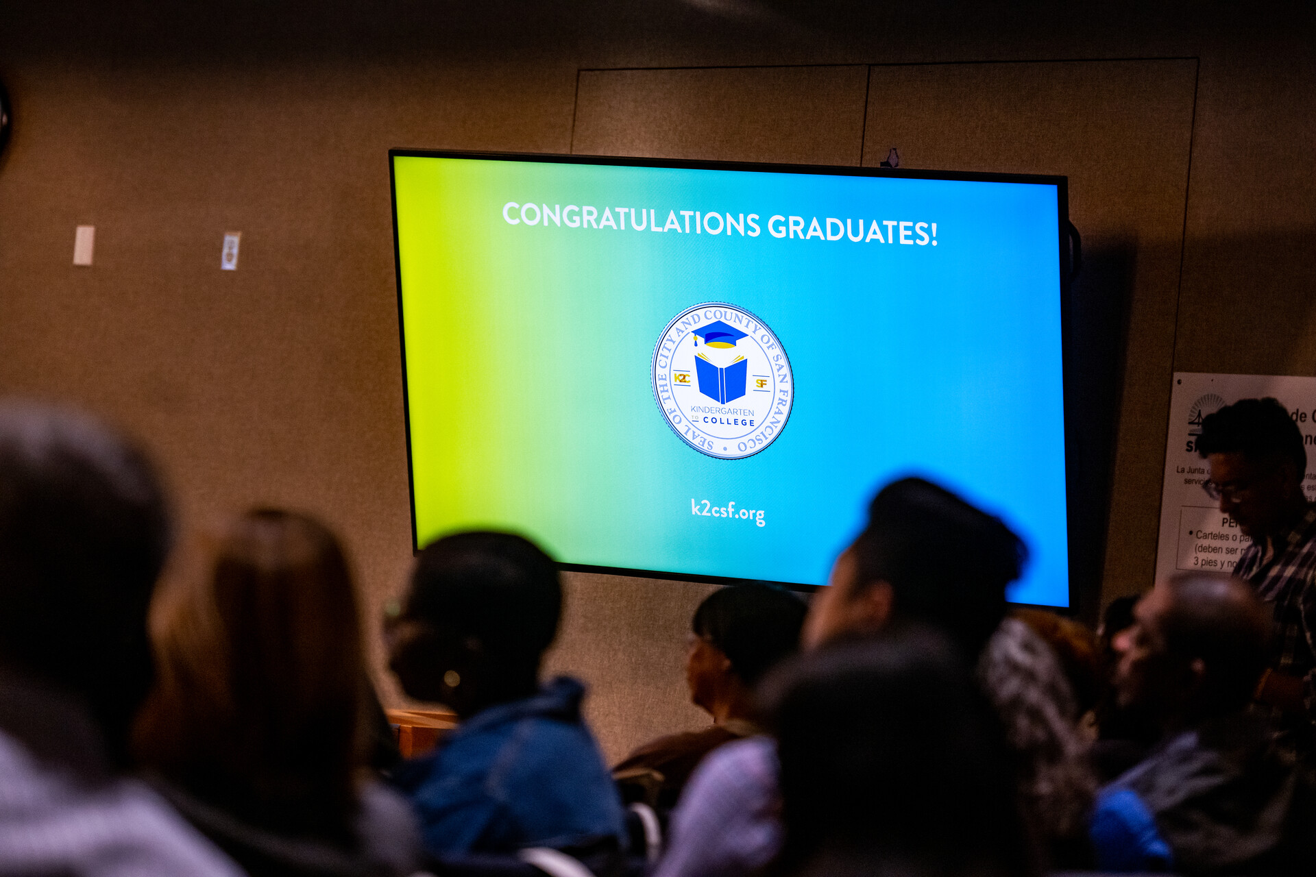 A crowd of people sit inside a large conference room and look away from the camera. Behind them, a large monitor reads out, "Congratulations graduates!"
