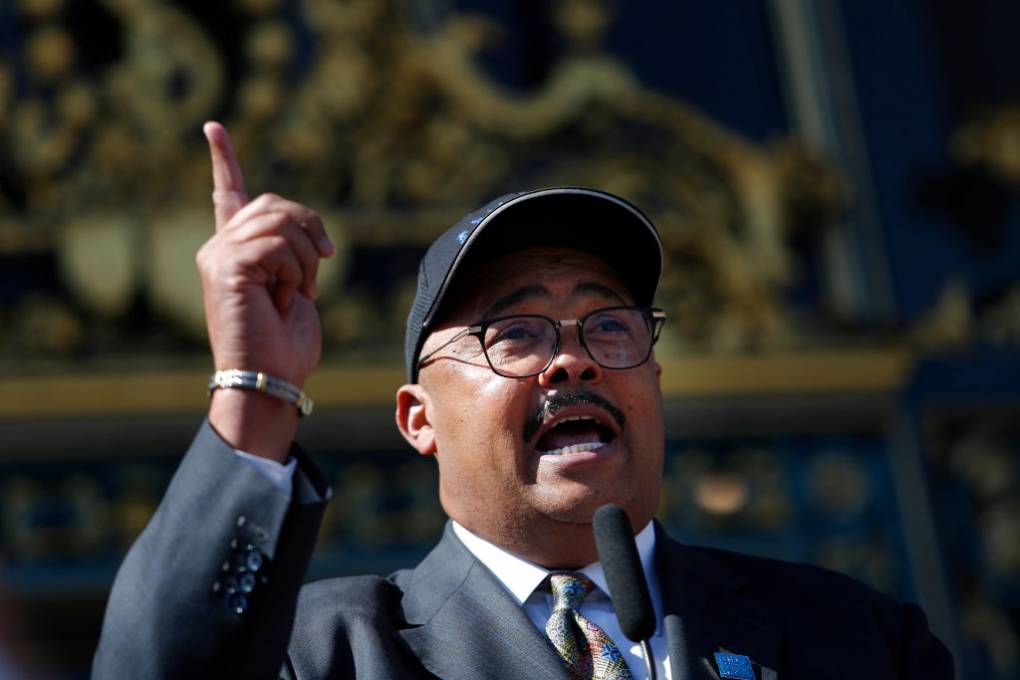 A man wearing business suit, glasses and a hat points up with his right hand outside in front of a microphone.