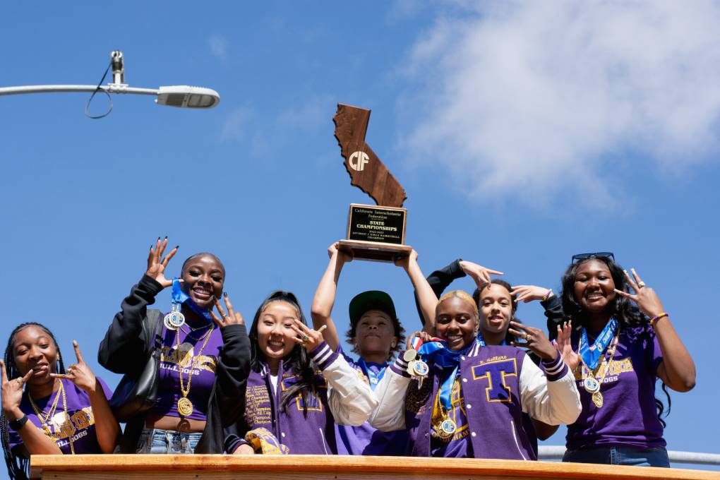 A group of young women wearing purple clothing stand on a bus waving with one person holding a trophy above their head.