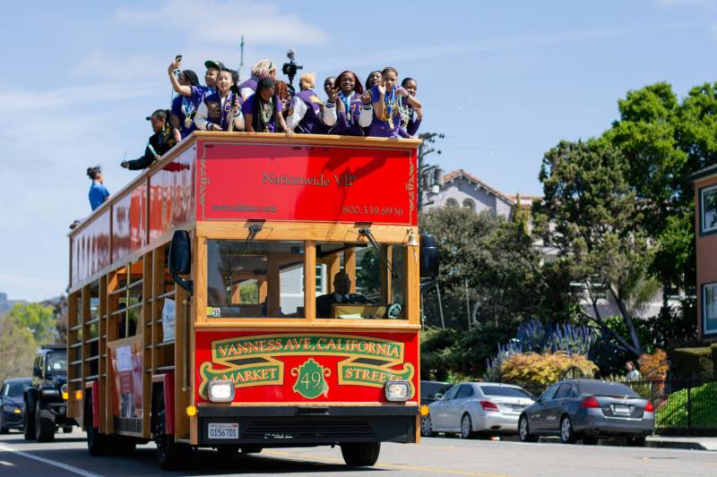 A group of teenage girls wearing purple and white stand on the top of a red double-decker bus driving down a city street, with parked cars and trees in the background.