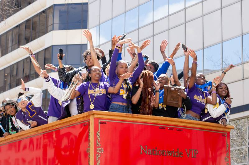 A group of female teenagers, all or mostly Black, stand with hands raised on the top of a red double decker bus, wearing purple T-shirts and varsity jackets and smiling.