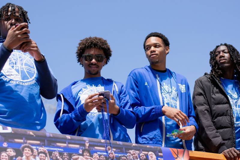 Four Black male teenagers wearing blue clothing look around and at their phones on top of a bus.