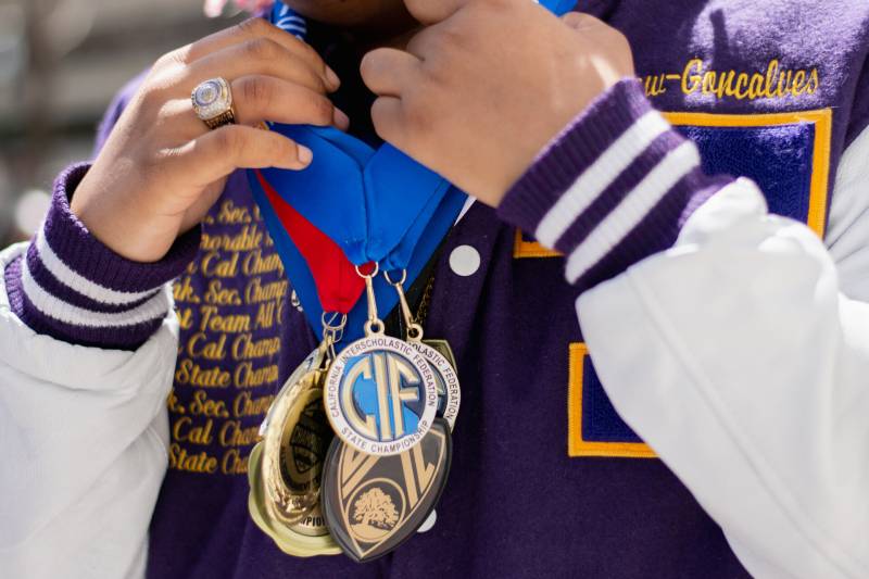 A closeup of multiple medals around a young woman's neck, which she holds with a hand that sports a championship ring, against a bright purple varsity jacket with white sleeves.