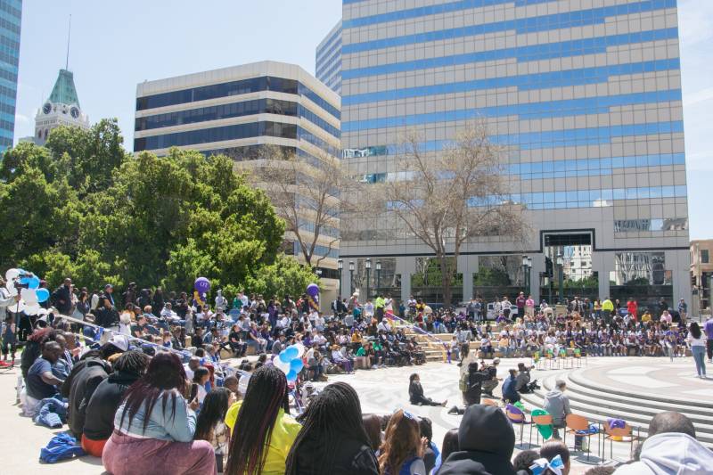 A large group of people of all ages sit in an outdoor amphitheater amid high-rise buildings.