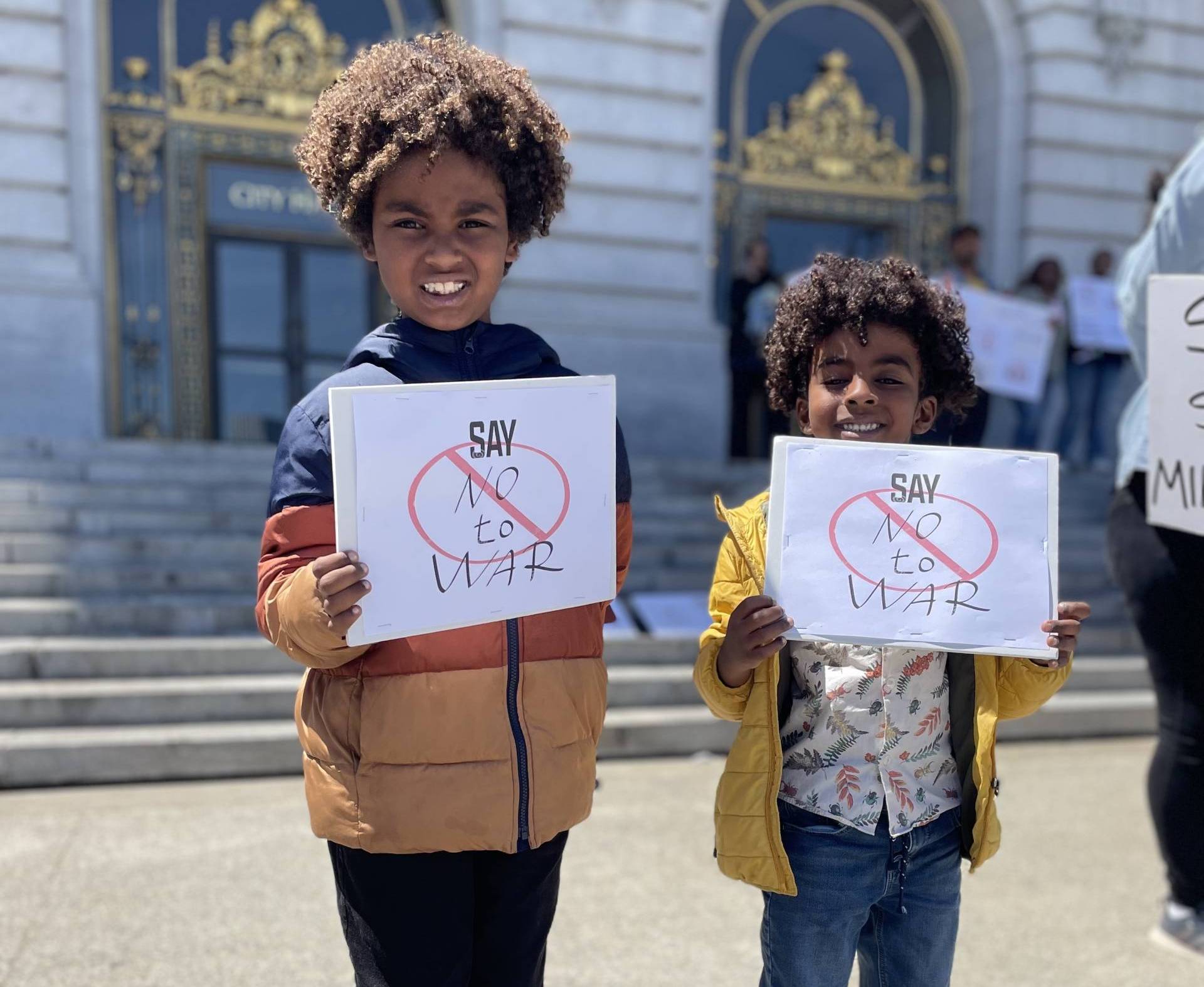 Two Sudanese American children, with long curly natural hair and colorful puffy jackets, hold signs outside SF City Hall.