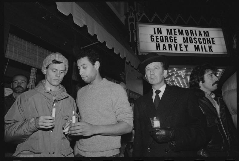 A white man and a Black man hold candles with another white man to the side and a sign above and behind them that says "In Memoriam George Moscone Harvey Milk"