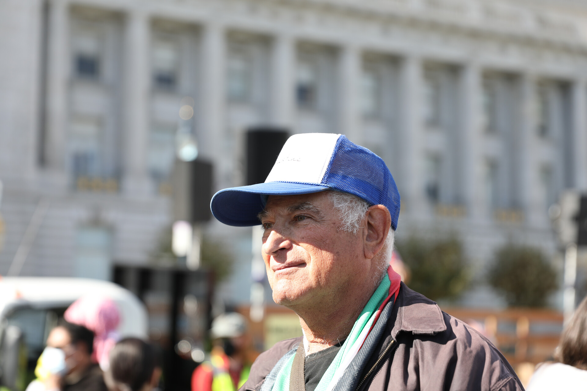 An older white man in a blue and white cap and a Palestinian flag draped around his neck looks away with a blurred crowd in the back.