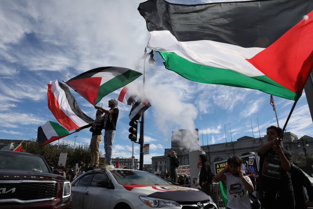 Men standing on cars wave Palestinian flags amid a crowd.