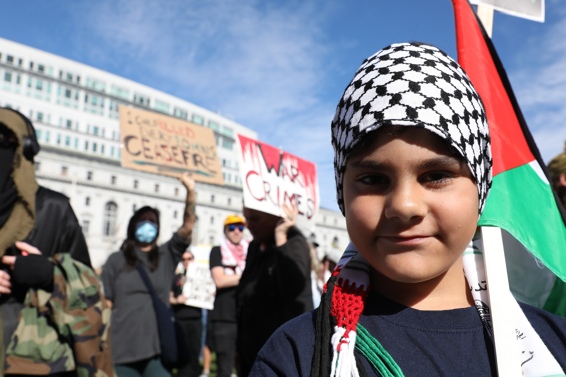 A young boy with a Palestinian flag looks into the camera as protesters chant and hold signs behind him.