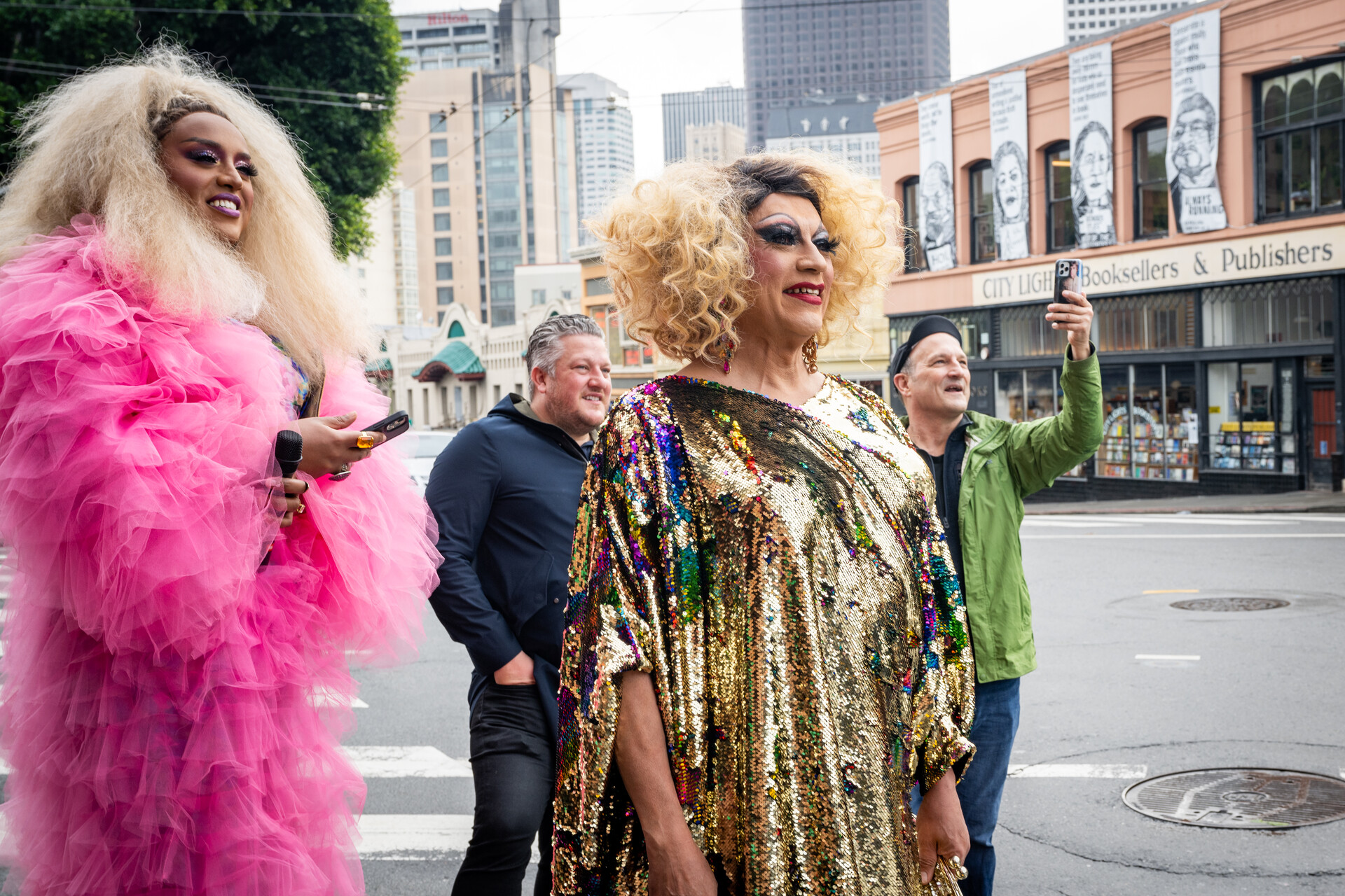 Two drag artists watch and smile on a street corner with some people in the background.