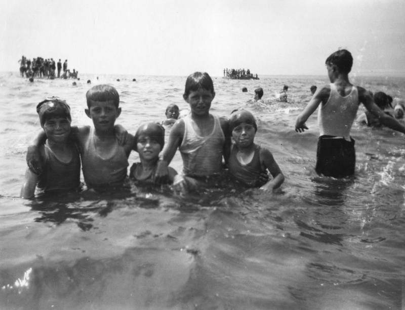 A line of very wet small children stand waist or chest-deep in the ocean, arms around each other warmly.