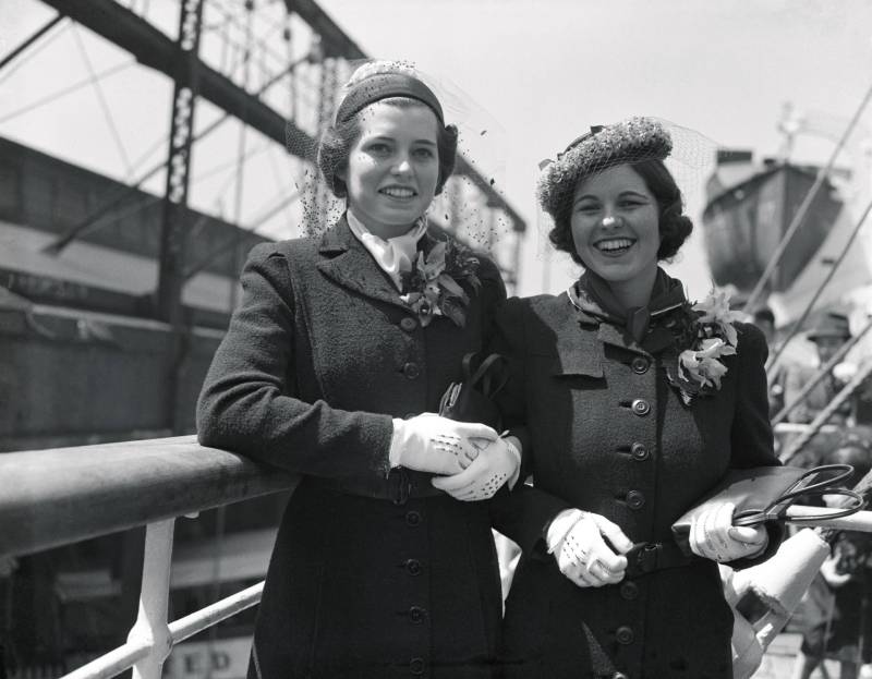 Two women in smart 1930s fitted coats and hats stand arm in arm smiling. They are both wearing white gloves.