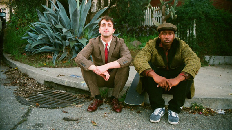 a white man in a suit and a Black man in a green jacket sit on a sidewalk looking at the camera