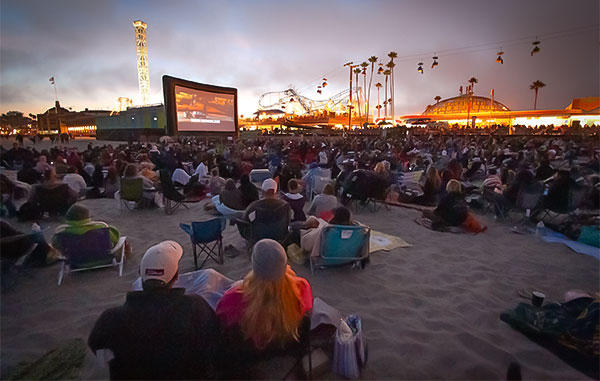 people watch a movie on an outdoor screen at the beach with a roller coaster in the background
