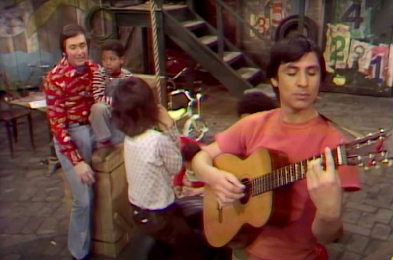 manin a flannel shirt plays with kids on Sesame Street while a Latino man in an orange shirt plays the acoustic guitar