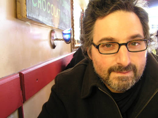A 30-something-year-old man with rectangular spectacles and a light beard sits at a diner table wearing a winter coat.