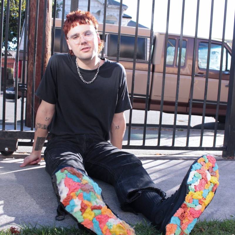 Musician Kevin Nichols sits on a sidewalk wearing all black clothing and rainbow shoes.