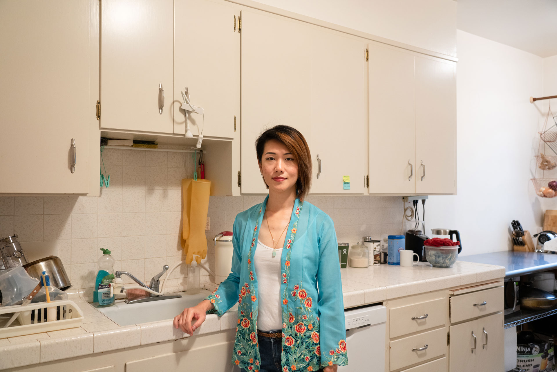 Woman in turquoise shirt leans one arm on the edge of sink, stands in her kitchen in front of cabinets and a long tiled counter.
