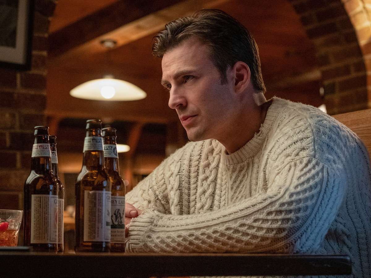 Chris Evans leans on a table in white cable knit sweater with four empty beer bottles in front of him.