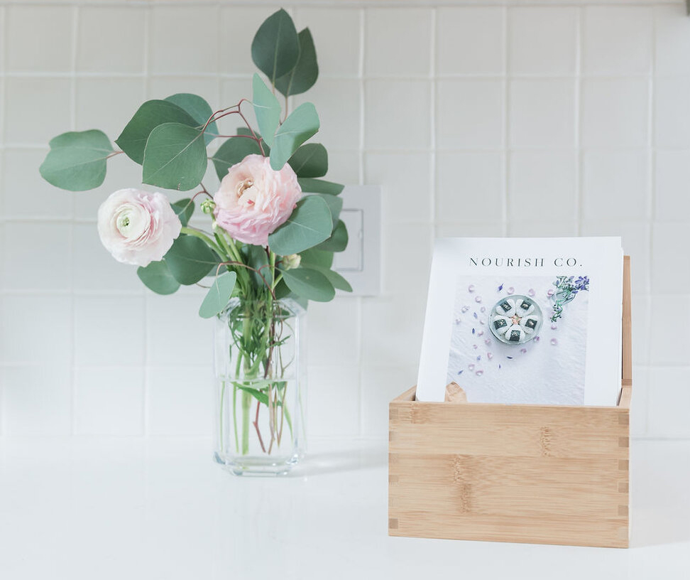 A cooking zine in a wooden box sits to the right of a glass vase of flowers on a white counter.