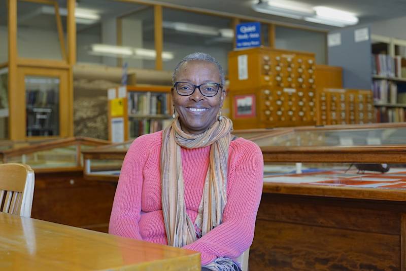 An older Black woman with short hair wears glasses, a pink sweater and a light scarf, smiling in a library