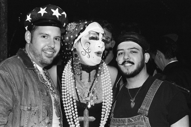 A black-and-white photo of a drag queen nun and two mustached men partying.