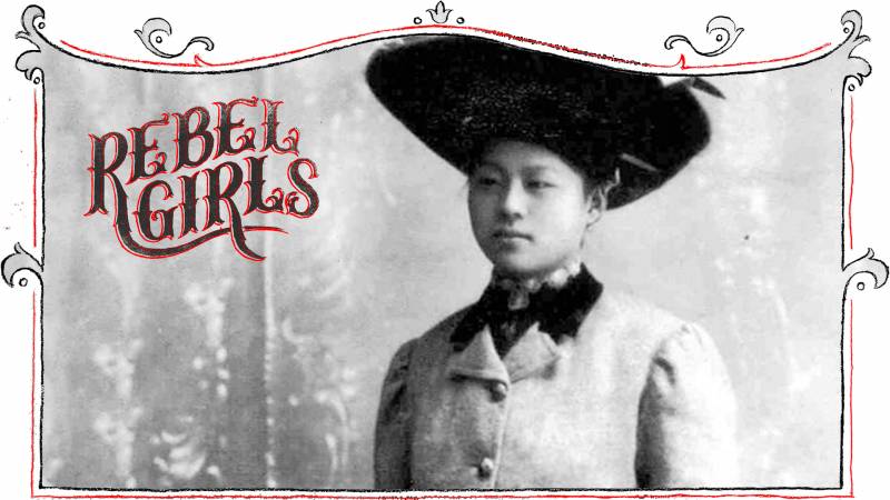A young woman of Chinese descent with a pretty round face poses for a portrait, wearing a large-brimmed black hat, smart suit jacket and lace choker.