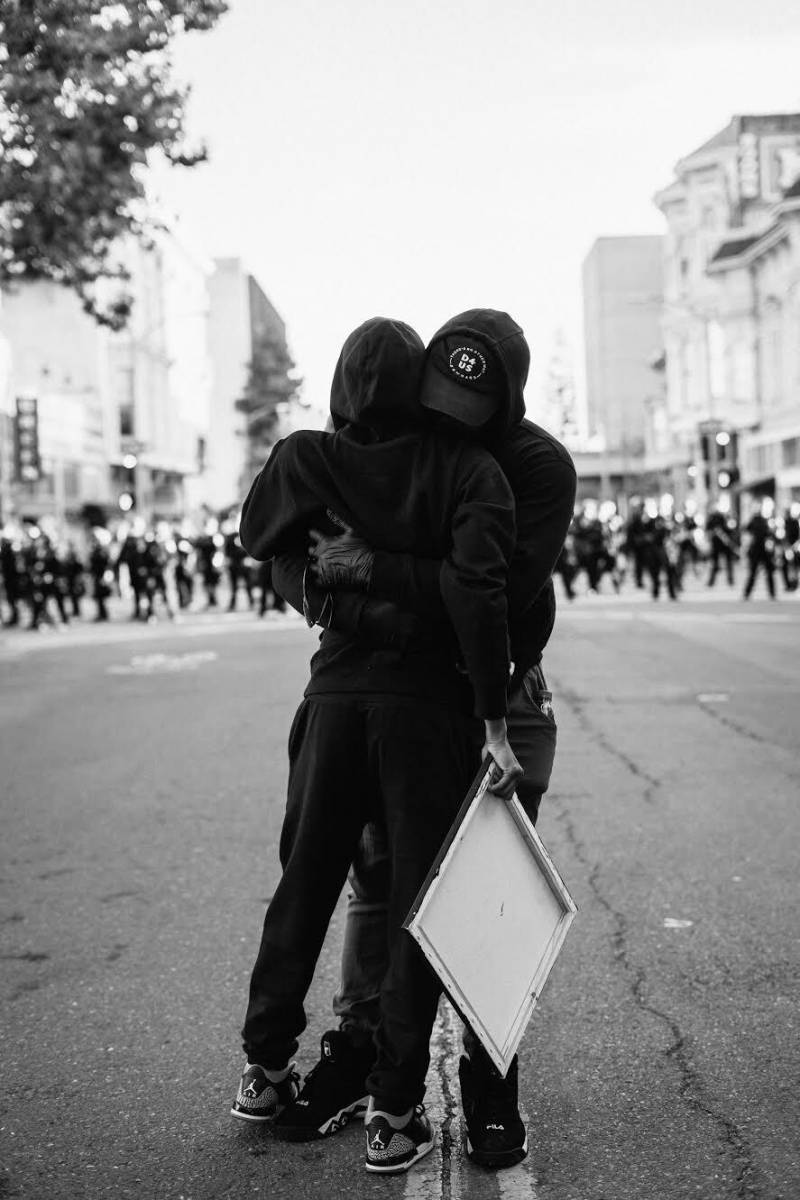 Two people wearing all black hug, behind them we see a police line in the distance.